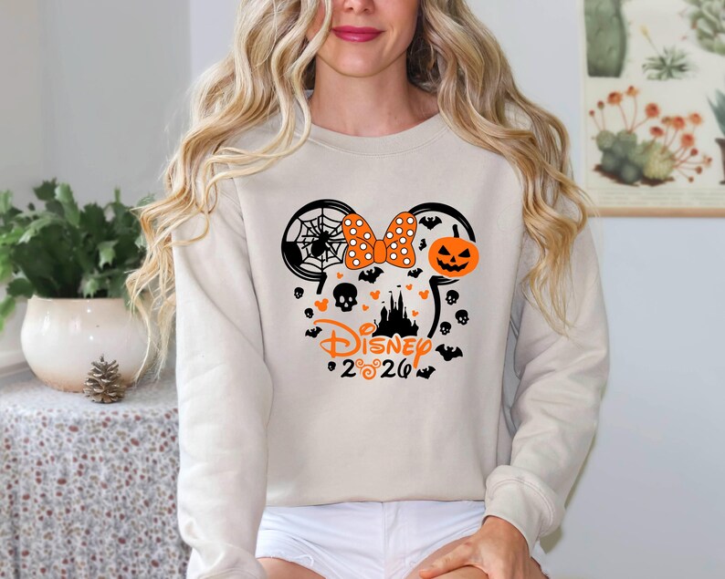 May include: A beige long-sleeve sweatshirt featuring a black and orange graphic of a Mickey Mouse silhouette filled with a spider web, a polka dot bow, a jack-o'-lantern, a castle, skulls, and bats, with the text Disney 2020 printed below.