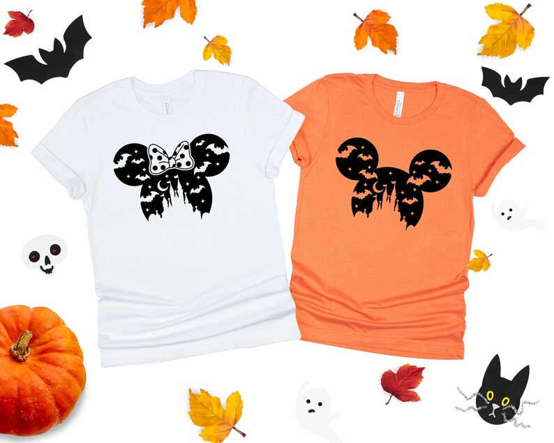 May include: Two t-shirts with a Halloween-themed Mickey Mouse silhouette design. One shirt is white and the other is orange. The design features Mickey Mouse ears with bats and a crescent moon.