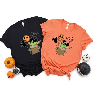 Pu&ograve; includere: Due t-shirt con un personaggio alieno verde che tiene palloncini arancioni e neri con orecchie di Topolino. L'alieno indossa una camicia marrone. Le t-shirt sono nere e arancioni.
