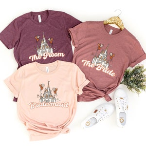 May include: Three t-shirts with a pink, maroon, and maroon color scheme. Each shirt features a white castle with orange balloons and text that reads "The Groom", "The Bride", and "Bridesmaid".