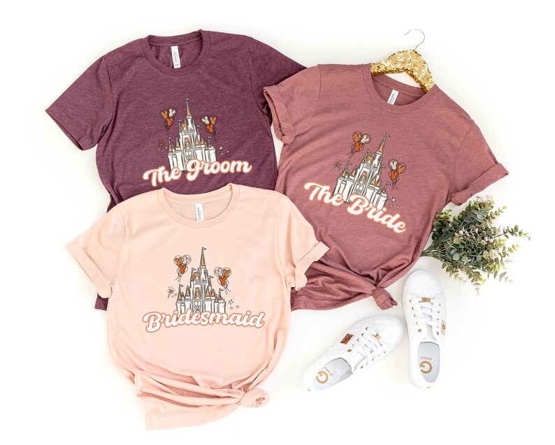 May include: Three t-shirts with a pink, maroon, and maroon color scheme. Each shirt features a white castle with orange balloons and text that reads "The Groom", "The Bride", and "Bridesmaid".