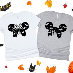 May include: Two t-shirts with a Halloween-themed Mickey Mouse design. One shirt is white with a black bow and the other is gray with a black Mickey Mouse head.