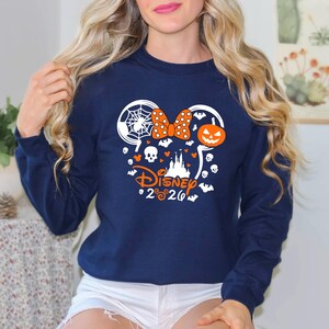 May include: A navy blue long-sleeve sweatshirt featuring a white and orange graphic of a mouse-ear silhouette filled with a spider web, a polka-dot bow, a jack-o'-lantern, skulls, bats, and a castle, with the text Disney 2026 printed below.