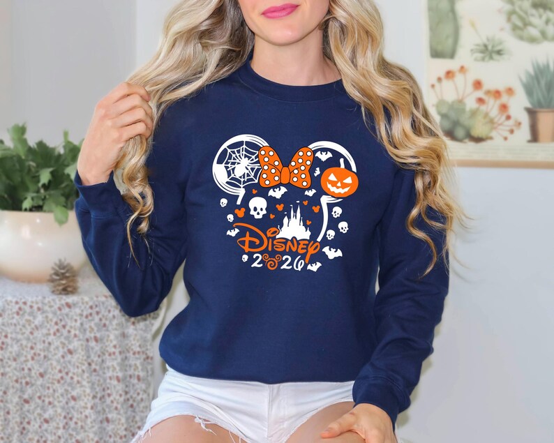 May include: A navy blue long-sleeve sweatshirt featuring a white and orange graphic of a mouse-ear silhouette filled with a spider web, a polka-dot bow, a jack-o'-lantern, skulls, bats, and a castle, with the text Disney 2026 printed below.