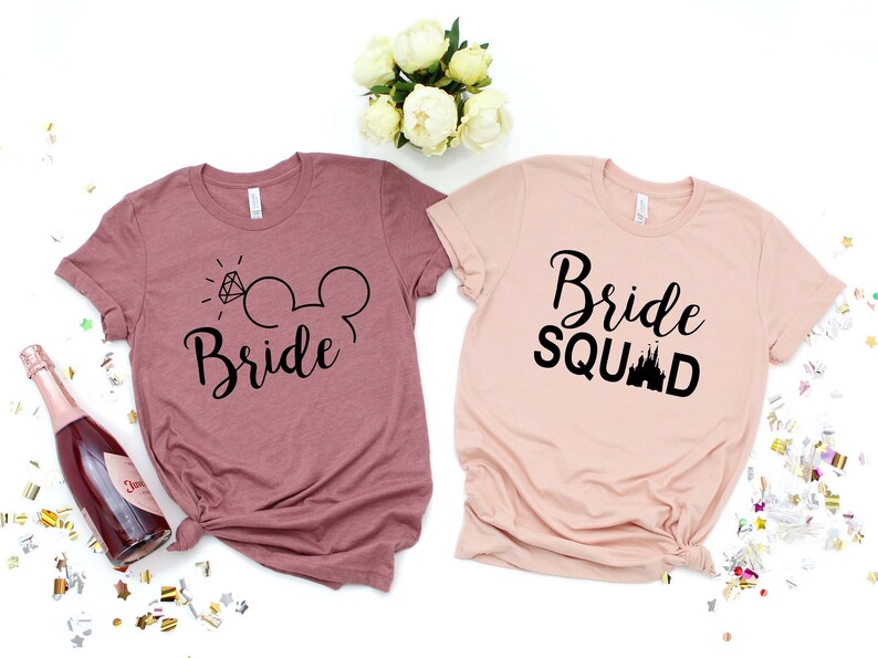 May include: Two t-shirts, one pink and one mauve, with a Disney theme. The pink shirt says "Bride Squad" with a black silhouette of Cinderella's castle. The mauve shirt says "Bride" with a black silhouette of Mickey Mouse ears and a diamond ring.