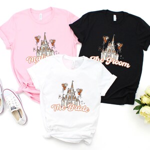 May include: Three t-shirts with a white castle graphic and text that reads "The Bride", "The Groom", and "The Bride". The shirts are pink, black, and white.