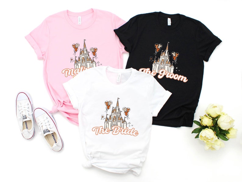 May include: Three t-shirts with a white castle graphic and text that reads "The Bride", "The Groom", and "The Bride". The shirts are pink, black, and white.