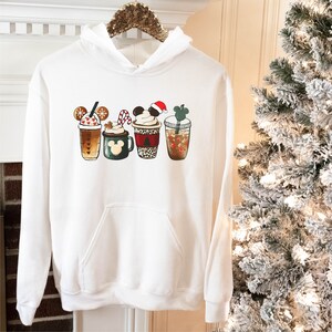 K&ouml;nnte beinhalten: Wei&szlig;er Kapuzenpullover mit einem Aufdruck von f&uuml;nf verschiedenen weihnachtlichen Kaffeegetr&auml;nken mit Mickey-Maus-Ohren. Die Getr&auml;nke umfassen einen Peppermint Mocha, einen Lebkuchen-Latte, eine hei&szlig;e Schokolade, einen Caramel Macchiato und einen Pumpkin Spice Latte.