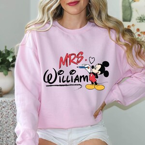 May include: Pink sweatshirt with a Mickey Mouse graphic and the text "MRS. WILLIAM" in black.