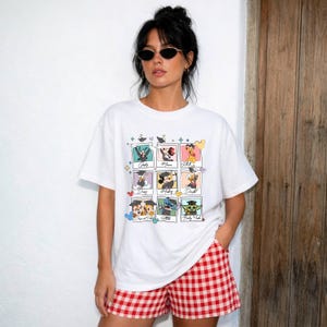 May include: White t-shirt featuring a collage of Disney characters in graduation caps. The characters include Goofy, Minnie Mouse, Donald Duck, Chip and Dale, Stitch, and Baby Yoda. The model is wearing red and white checkered shorts.
