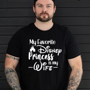 May include: Black t-shirt with white text that reads "My Favorite Disney Princess is my Wife." The text includes a castle graphic and the word "Disney" in a stylized font. The shirt is a crew neck.