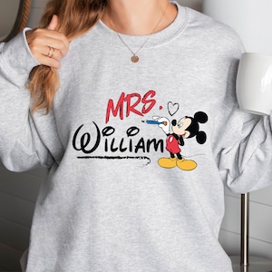 May include: Gray sweatshirt with a red and black Mickey Mouse design. The text "MRS. William" is written in black with a heart next to the word "MRS."