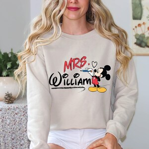 May include: A beige sweatshirt with a Mickey Mouse graphic and the text "MRS. William" in black and red.
