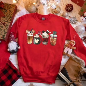 K&ouml;nnte beinhalten: Roter Weihnachtspullover mit einem Aufdruck von f&uuml;nf verschiedenen festlichen Kaffeetassen. Die Tassen sind mit Mickey-Mouse-Ohren, Zuckerstangen und anderen festlichen Details verziert.
