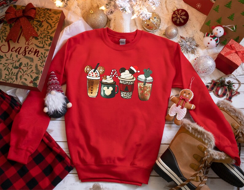 K&ouml;nnte beinhalten: Roter Weihnachtspullover mit einem Aufdruck von f&uuml;nf verschiedenen festlichen Kaffeetassen. Die Tassen sind mit Mickey-Mouse-Ohren, Zuckerstangen und anderen festlichen Details verziert.