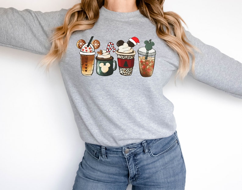 K&ouml;nnte beinhalten: Ein graues Sweatshirt mit einem festlichen Design aus f&uuml;nf verschiedenen festlichen Kaffeetassen mit Mickey-Maus-Ohren. Die Tassen sind mit Schneeflocken, Zuckerstangen und Weihnachtsb&auml;umen verziert.