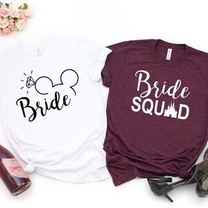 May include: Two white t-shirts with black text. One shirt says "Bride" with a diamond ring and Mickey Mouse ears. The other shirt says "Bride Squad" with a white castle graphic.