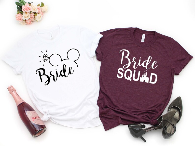 May include: Two white t-shirts with black text. One shirt says "Bride" with a diamond ring and Mickey Mouse ears. The other shirt says "Bride Squad" with a white castle graphic.