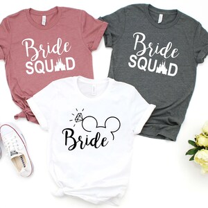 May include: Three t-shirts with the text "Bride Squad" and a Disney castle graphic. The white t-shirt has a diamond ring and Mickey Mouse ears with the text "Bride".
