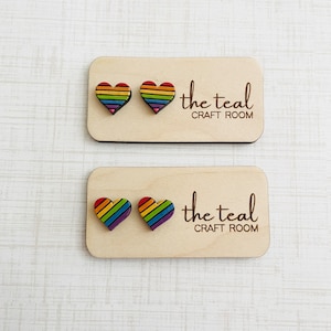 May include: Two pairs of rainbow heart shaped wooden earrings on a wooden display card with the text "the teal craft room" printed on it.
