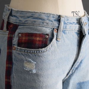 K&ouml;nnte beinhalten: Hellblaue Jeans mit einer ausgefransten Tasche und einem rot-schwarz karierten Akzent an der Seite und der Tasche. Die Jeans haben ein klassisches F&uuml;nf-Taschen-Design und einen Knopf- und Rei&szlig;verschluss. Die Jeans werden auf einer Schaufensterpuppe pr&auml;sentiert.