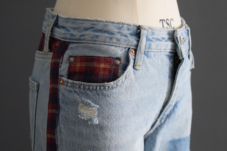 K&ouml;nnte beinhalten: Hellblaue Jeans mit einer ausgefransten Tasche und einem rot-schwarz karierten Akzent an der Seite und der Tasche. Die Jeans haben ein klassisches F&uuml;nf-Taschen-Design und einen Knopf- und Rei&szlig;verschluss. Die Jeans werden auf einer Schaufensterpuppe pr&auml;sentiert.