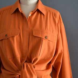 May include: Orange long-sleeved shirt with two front pockets and button closures. The shirt is tied at the waist, and is displayed on a mannequin. The shirt is paired with blue jeans.