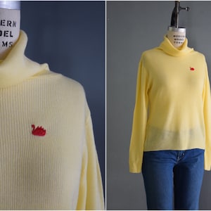 May include: A pale yellow turtleneck sweater with a small red embroidered swan on the front. The sweater is on a mannequin.