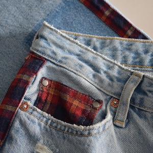 K&ouml;nnte beinhalten: Hellblaue Jeans mit einem rot, schwarz und gold karierten Akzent an der Seite und der Tasche. Die Jeans haben einen Used-Look mit verwaschener Optik und sichtbaren N&auml;hten. Die Tasche hat ein Karomuster.