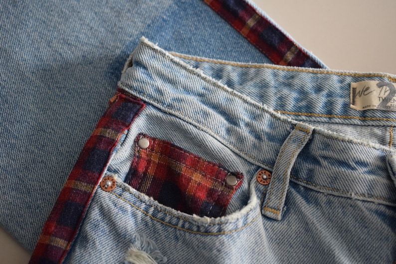 K&ouml;nnte beinhalten: Hellblaue Jeans mit einem rot, schwarz und gold karierten Akzent an der Seite und der Tasche. Die Jeans haben einen Used-Look mit verwaschener Optik und sichtbaren N&auml;hten. Die Tasche hat ein Karomuster.
