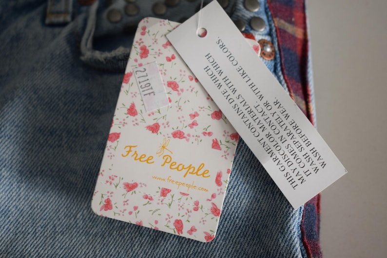 K&ouml;nnte beinhalten: Nahaufnahme einer Jeans aus Denim mit einem floralen Etikett von Free People. Das Etikett hat rosa Rosen auf wei&szlig;em Grund und den Text "Free People". Ein zweites Etikett enth&auml;lt Waschanweisungen.