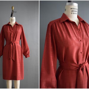 Puede incluir: Vestido vintage color óxido con cuello, mangas largas y cinturón anudado. El vestido presenta un sutil estampado de cuadros y se exhibe en un maniquí. El vestido mide aproximadamente 102 cm de largo.