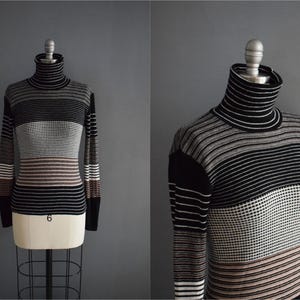 May include: A vintage turtleneck sweater with horizontal stripes in black, white, brown, and gray. The long-sleeved sweater is displayed on a mannequin against a gray backdrop. The turtleneck is black and white striped.