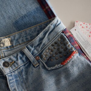 K&ouml;nnte beinhalten: Hellblaue Jeans mit Used-Look, mit einer verzierten Tasche und einem rot-blau karierten Akzent. Die Jeans haben einen Knopfverschluss und ein "The 26"-Etikett. Ein "Free People"-Etikett ist angebracht.