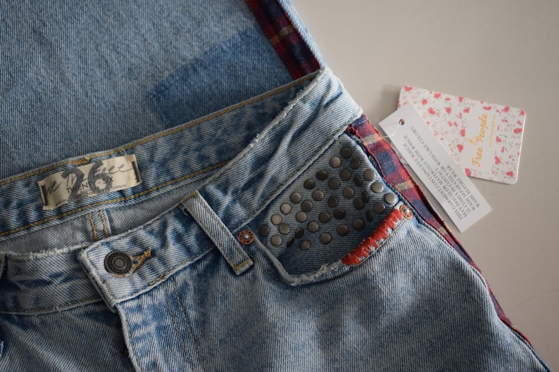K&ouml;nnte beinhalten: Hellblaue Jeans mit Used-Look, mit einer verzierten Tasche und einem rot-blau karierten Akzent. Die Jeans haben einen Knopfverschluss und ein "The 26"-Etikett. Ein "Free People"-Etikett ist angebracht.