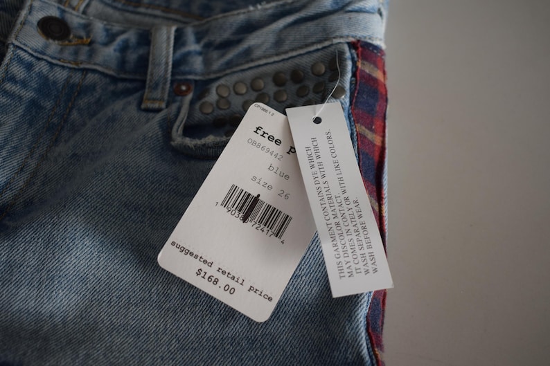 K&ouml;nnte beinhalten: Hellblaue Jeans mit einem rot-blau karierten Seitenakzent. Die Jeans hat Metallnieten und ein Etikett mit "free people" und Gr&ouml;&szlig;e 26. Der empfohlene Verkaufspreis betr&auml;gt 168,00 $.