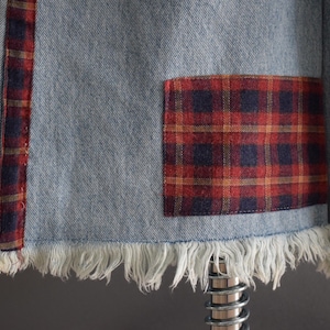 K&ouml;nnte beinhalten: Nahaufnahme eines Jeanskleidungsst&uuml;cks mit ausgefranstem Saum. Das Kleidungsst&uuml;ck hat einen rot-marine karierten Aufn&auml;her und einen rot-marine karierten Streifen. Der Denim ist hellblau.