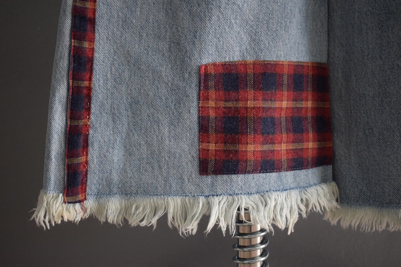 K&ouml;nnte beinhalten: Nahaufnahme eines Jeanskleidungsst&uuml;cks mit ausgefranstem Saum. Das Kleidungsst&uuml;ck hat einen rot-marine karierten Aufn&auml;her und einen rot-marine karierten Streifen. Der Denim ist hellblau.