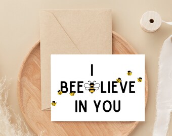 May include: White card with black text that reads "I BEElieve in you". A bee illustration is in the middle of the text. There are small bee illustrations scattered around the card.