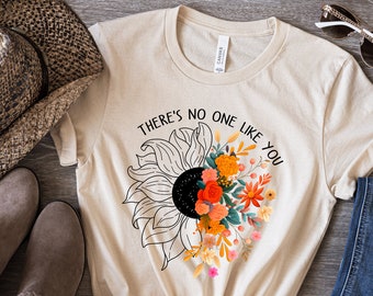 May include: A beige t-shirt with a half-sunflower design, black center, and orange, red, and yellow floral arrangement. The text "THERE'S NO ONE LIKE YOU" is above the sunflower. The shirt is knotted at the waist.