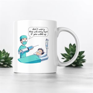 Puede incluir: Taza de cerámica blanca con una ilustración de dibujos animados de un médico que le da una inyección a un paciente. El texto en la taza dice "no te preocupes, esto solo dolerá si te despiertas".