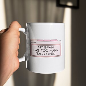 May include: White ceramic mug with a pink and black design. The design features a browser window with the text "My brain has too many tabs open."