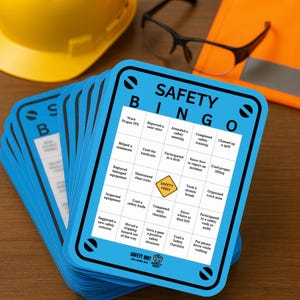 Puede incluir: Una pila de tarjetas azules de Bingo de seguridad con bordes negros. Las tarjetas tienen una cuadrícula de acciones relacionadas con la seguridad y las palabras "SAFETY BINGO" en la parte superior. Un casco amarillo, gafas de seguridad y una carpeta naranja están en el fondo.