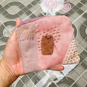 May include: A pink and mauve fabric pouch with a zipper closure. The pouch features a brown, cartoon-like character applique and gold-colored stitched details. The pouch is held in a hand.