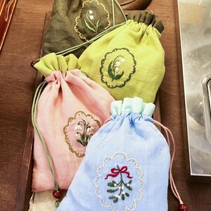 Puede incluir: Una colección de bolsas con cordón en varios colores: verde oliva, verde claro, rosa y azul. Cada bolsa presenta un diseño floral bordado dentro de un borde festoneado. Las bolsas tienen cordones a juego y se exhiben en una bandeja de madera.