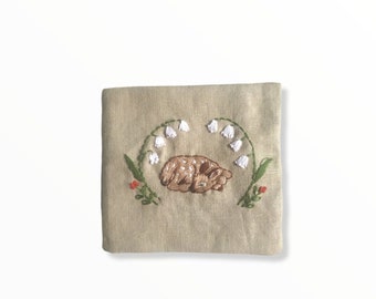 May include: Embroidered beige square featuring a sleeping fawn, encircled by white lily of the valley flowers and green stems. Small red flowers accent the design. The embroidery is on a beige linen-like fabric.