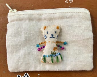 Puede incluir: Pequeño monedero de lino blanquecino con cierre de cremallera. Una muñeca de gato hecha a mano con cuentas azules, rosas y verdes está unida. Una ilustración de cara de gato y pez está en la esquina inferior izquierda.