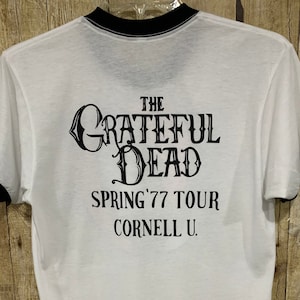 May include: White t-shirt with black trim around the collar and sleeves. The back of the shirt features the text "THE GRATEFUL DEAD SPRING '77 TOUR CORNELL U." in black lettering. The shirt is made of a lightweight material.