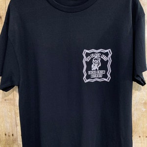 以下が含まれることがあります： クルーネックと半袖の黒いTシャツ。左胸には、スカルのデザインで「The Black Keys Local Crew Detroit」と書かれた白いグラフィックがあります。シャツは白いハンガーに掛けられ、木製の背景にあります。