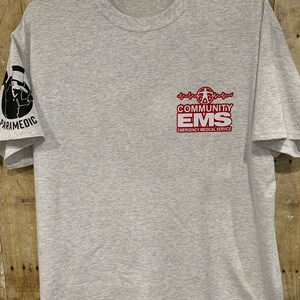 Pode incluir: Camiseta cinza claro com a inscrição "COMMUNITY EMS EMERGENCY MEDICAL SERVICE" em vermelho no peito. A manga esquerda tem um desenho preto de uma pessoa com um chapéu e a palavra "PARAMEDIC".